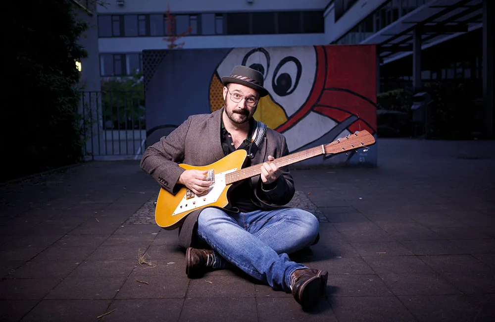 Sebastian Bogensperger mit Hut und E-Gitarre, im Schneidersitz vor einer Wand mit Vogel-Graffiti, Aufnahme zur blauen Stunde