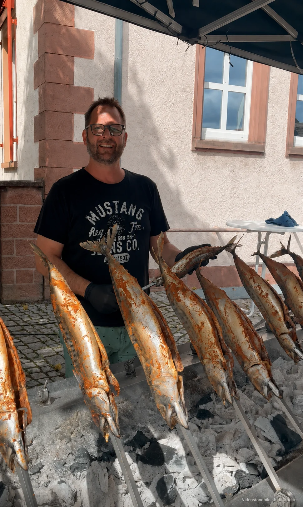 Fische auf Holzspießen über Holzkohle, dahinter ein Mann am Grill (Videostandbild · KI-bearbeitet)